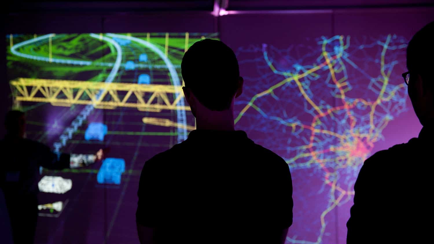 A person silhouetted against projector screens showing a 3d wireframe of vehicles on a road, and an overhead heatmap of roads in a large city